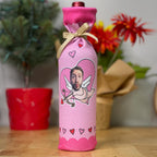 A pink fabric wine bottle cover with a custom printed cartoon cupid design featuring a man's face, tied with a beige ribbon, standing on a wooden table with blurred potted plants and flowers in the background.
